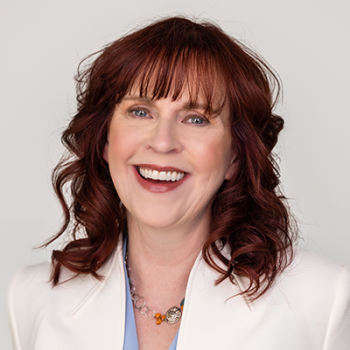 A smiling woman with shoulder-length reddish-brown hair wearing a light blue top and a white blazer.