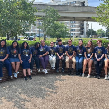 A group of fourteen young people wearing matching dark blue t-shirts with a gold circular graphic sit in a row on a low brick wall.