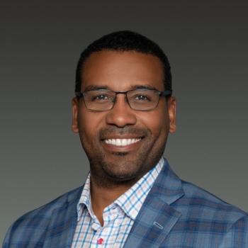 A smiling man wearing glasses and a plaid blazer over a light-colored button-up shirt.