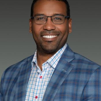A smiling man wearing glasses and a plaid blazer over a light-colored button-up shirt.