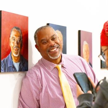 A smiling man in a pink button-down shirt and yellow tie stands in a gallery next to a portrait of a man, while a person in the foreground photographs him with a phone.