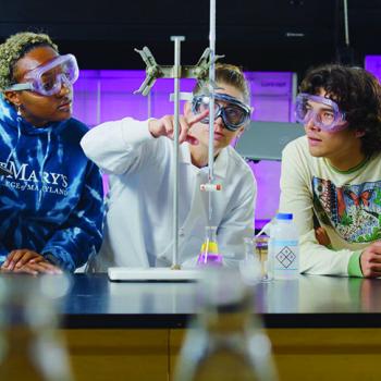 Three students wearing safety goggles observe a science experiment with beakers and lab equipment in a classroom setting.