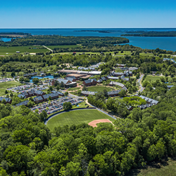 Aerial view of a campus with multiple buildings and a baseball field surrounded by trees, situated near a large body of water.