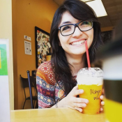 Caitlin smiling with glasses on holding a mango smoothie in a coffeehouse.
