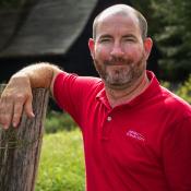 A man with a red shirt stands next to a tree outside