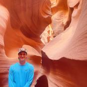 Matthew, wearing blue long sleeve shirt and hat, smiling and standing inside a slot canyon in Northern Arizona.