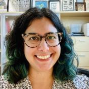 Me, Liz Leininger, looking at the camera and smiling. I have dark brown hair with parts dyed teal, wear tortise-shell pattern glasses and am wearing a white shirt with black leaf pattern
