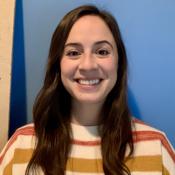 Rachel Heiss is smiling at the camera, wearing an orange, yellow and white striped sweater and standing in front of a blue wall.