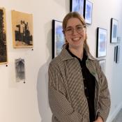 A woman smiles while standing in front of her artwork in a gallery.