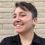A woman with short brown hair, wearing a black button-down shirt, stands in front of a brick wall. She is smiling, wearing glasses on top of her head. 