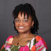 Alise Maxie is wearing a vibrant pink and green dress paired with gold wire glasses.