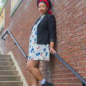 Black woman with red head wrap, natural black hair, wearing a black blazer, a white dress with red and blue flowers, and black boots