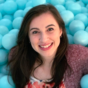 A person with long dark hair smiles while lying in a pit filled with light blue plastic balls. They are wearing a floral shirt and a maroon cardigan.