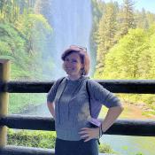 Sarah Grzyb (Sustainability Fellow) standing with hands on their hips and smiling in front of a waterfall.
