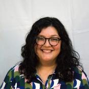A woman with curly black hair and round brown glasses smiles at the camera.