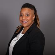 Smiling person with dreadlocks, wearing a black blazer over a white polka-dotted shirt, posing against a gray background.