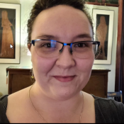 A person with glasses smiles at the camera. They have dark hair pulled back and are wearing a necklace. Behind them are framed artworks on the wall.