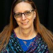 A woman with long auburn hair and glasses, wearing a paisley top with blue, smiles against a dark background.