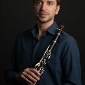 A person with short hair holds a clarinet against a dark background. They are wearing a dark blue shirt and a bracelet, looking at the camera with a neutral expression.