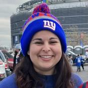 A person smiles outdoors wearing a blue and red beanie with a "NY" logo. In the background, there's a stadium and parked cars. It's a cloudy day, and the person is dressed warmly.