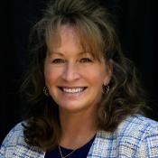 Smiling person with wavy hair wearing a blue and white jacket and a gold necklace against a dark background.