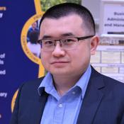 A person wearing glasses and a blue collared shirt under a dark blazer is standing in front of educational posters about business administration. The posters have text about business training and skills development.