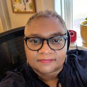Person with short gray hair and glasses sitting indoors in front of a window, wearing a dark top. There's a potted plant and small red item on the window sill.