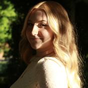 A young woman with long blonde hair and a white lace top stands outdoors, partially in sunlight and shadow, looking over her shoulder with a slight smile.