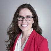 Woman with long brown hair and glasses wearing a red blazer and white blouse smiles at the camera against a plain background.