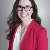 Woman with long brown hair and glasses wearing a red blazer and white blouse smiles at the camera against a plain background.