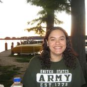 A woman with curly hair smiles at the camera. She is wearing a green "United States Army" t-shirt. Kayaks and boats are visible in the background near a lakeside at sunset.