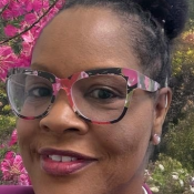 Woman wearing large, colorful eyeglasses and a purple blazer, standing outdoors in front of blooming pink flowers and greenery.
