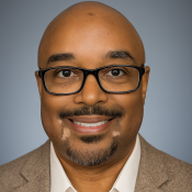 A man with a shaved head, glasses, and a goatee, wearing a tan blazer and white shirt, smiling at the camera against a plain background.