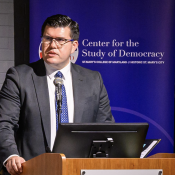A man in a suit speaks at a podium with a microphone, in front of a banner for the Center for the Study of Democracy at St. Mary’s College of Maryland.