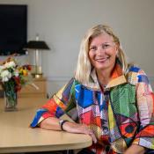 A woman with blonde hair and a colorful jacket sits at a conference table, smiling, with a bouquet of flowers and a lamp in the background.