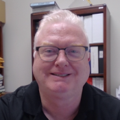 A man with short, light hair and glasses, wearing a black shirt, sits in an office in front of a shelf with binders and papers.