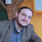 A person with short brown hair, glasses, and a beard, wearing a brown blazer and gray shirt, sits indoors near a wooden beam and a yellow wall.
