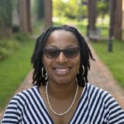 A woman wearing glasses, a striped dress, a pearl necklace, and hoop earrings smiles outdoors on a brick path with greenery in the background.