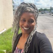 A woman with long, curly gray hair smiles outdoors near a beige building and a parking lot on a sunny day.