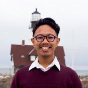 A person wearing glasses and a maroon sweater stands outdoors, smiling, with a house and lighthouse in the background.