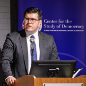 A man in a suit speaks at a podium with a microphone in front of a sign for the Center for the Study of Democracy at St. Mary's College of Maryland.