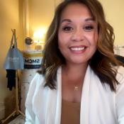 A woman with shoulder-length brown hair and a white blazer smiles at the camera in an office setting with a lamp, coat rack, and SMCM sign in the background.