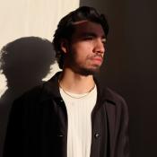 Photo of Instructor Joey Antico -- a man dark hair and a beard stands wearing a white sweater, black jacket and a thin gold necklace