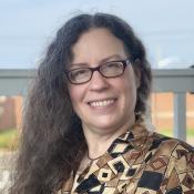 A person with long curly hair and glasses smiles at the camera, sitting outdoors. They are wearing a patterned shirt, and a fence is visible in the background.