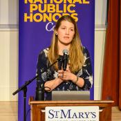 A woman stands at a podium labeled "St. Mary's College of Maryland," speaking into a microphone in front of a "National Public Honors College" banner.
