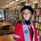 Person in academic regalia stands and smiles in a library, with bookshelves and study tables visible in the background.