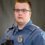  A Public Safety Officer in uniform with a badge, body camera, and radio headset, standing against a plain, neutral background."