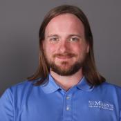 A person with long brown hair and a beard smiles at the camera, wearing a blue St. Mary’s College of Maryland polo shirt against a plain gray background.
