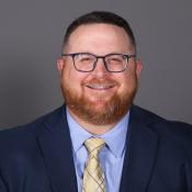A man with short hair, a beard, and glasses, wearing a navy suit, light blue shirt, and yellow plaid tie, is smiling in front of a plain gray background.