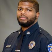 A man in a police uniform with badges and patches poses for a formal portrait against a plain gray background.